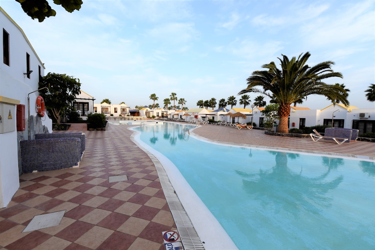 The image showcases a large swimming pool surrounded by palm trees and shaded areas. The pool features a winding design with inviting turquoise water and is bordered by tiled walkways. Sun loungers are positioned alongside the pool, offering space for relaxation.