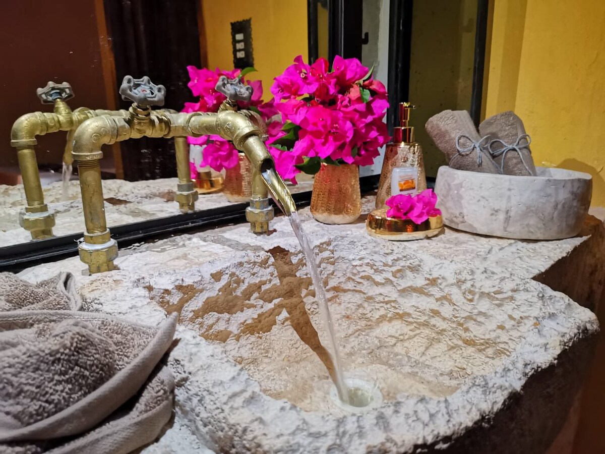 A rustic bathroom sink crafted from stone is featured, with water flowing gracefully from decorative brass faucets. Nearby, a small arrangement of vibrant pink flowers adds a touch of color, complemented by neatly arranged towels and spa essentials on a textured surface.