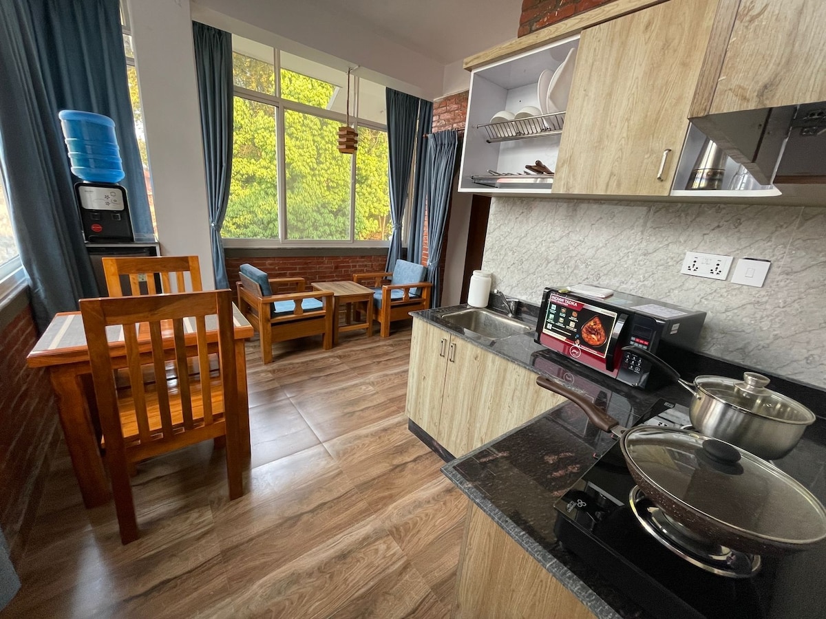 A functional kitchen area features wooden furniture, including a dining table and two chairs. A microwave and stovetop are visible on the countertop, alongside a cooking pot. Large windows provide natural light, framed by blue curtains, while a water dispenser is positioned near the entrance.