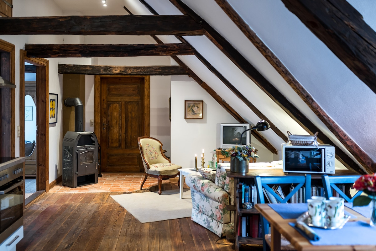 A cozy living area is showcased within an attic-like space featuring sloped wooden beams. A floral-patterned sofa is positioned near a small table, complemented by ambient candlelight. A vintage-style chair adds character, while a modern microwave and sturdy bookshelf complete the practical setting.