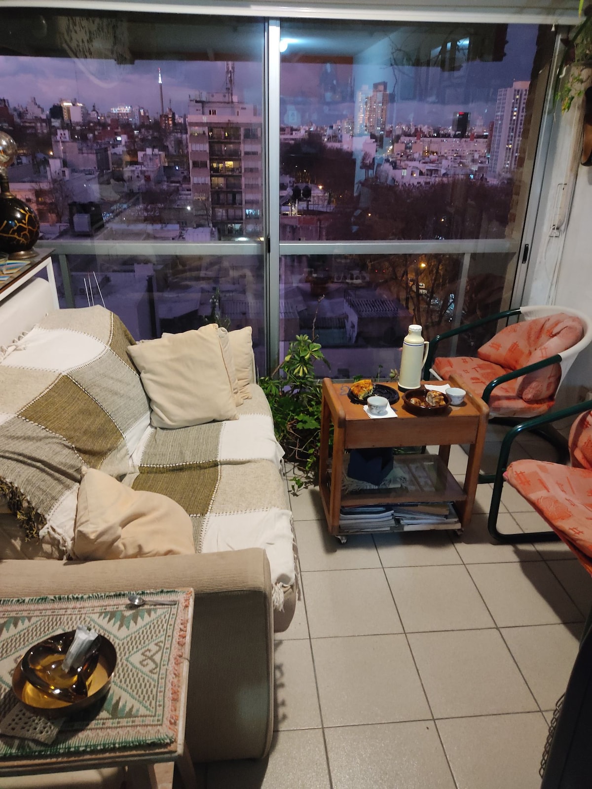 A cozy sitting area is featured with a light-colored sofa adorned with cushions. A small wooden table holds refreshments, surrounded by two chairs. The large windows provide a view of the city skyline during dusk, enhancing the inviting ambiance.