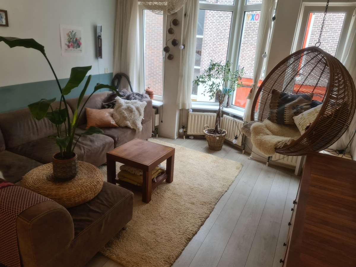 A cozy living area features a spacious sofa adorned with cushions, complemented by a wooden coffee table and a soft area rug. A hanging chair provides additional seating, while large windows allow natural light to brighten the space. Potted plants add a touch of greenery.