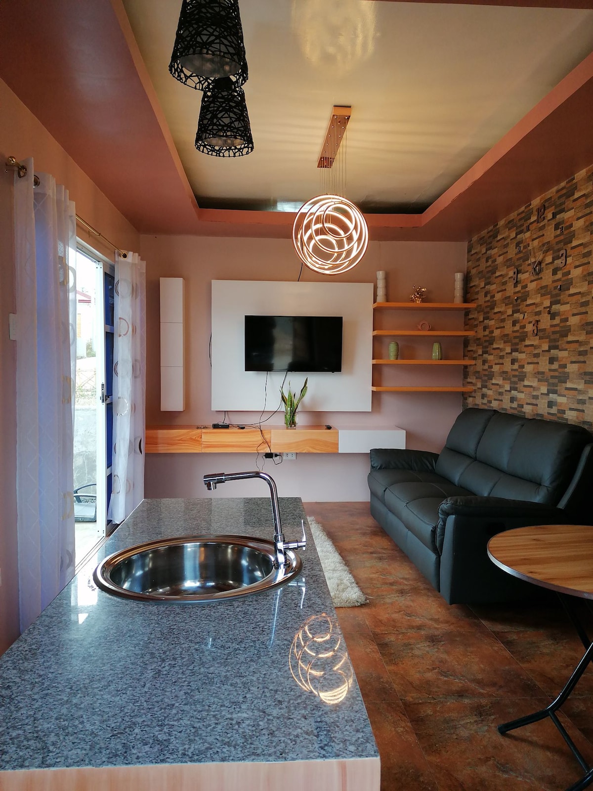The living area features a sleek dark sofa and a wall-mounted television. Light wood shelving holds decorative items. A round dining table is visible, along with a kitchen counter that includes a sink, all bathed in soft natural light from the adjacent window.