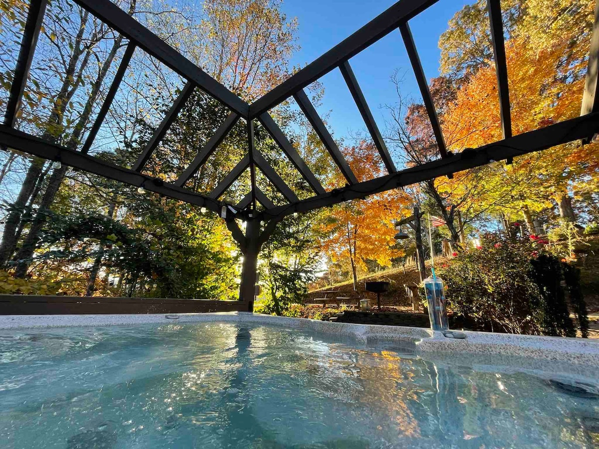 An outdoor hot tub is framed by a wooden pergola, surrounded by vibrant autumn foliage. Sunlight filters through the trees, creating a serene ambiance. The water surface reflects the clear sky, inviting relaxation and enjoyment of nature.