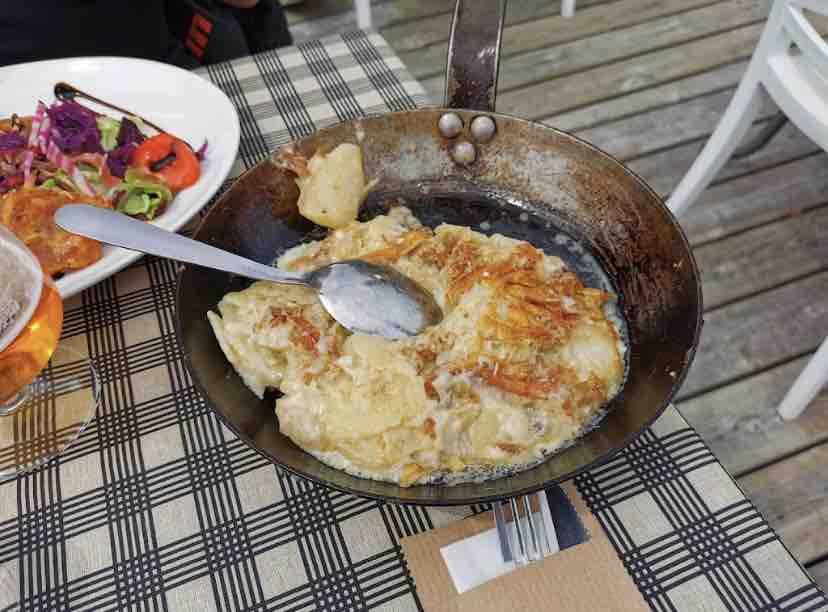 A portion of baked dish is served in a round metal pan with a shiny finish. The top appears golden and crispy, suggesting a warm, hearty meal. A spoon rests inside the pan, and a faint wooden surface is visible underneath, accompanied by a patterned tabletop.