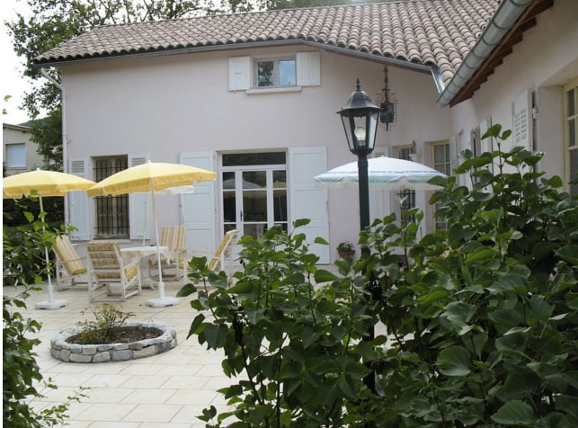 A charming outdoor patio area features yellow and blue umbrellas over several seating arrangements. A stone fire pit is visible in the center, surrounded by greenery. The light-colored walls of the nearby house and large French doors create a welcoming environment.