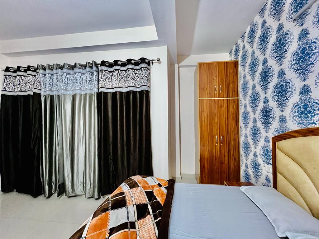 A spacious bedroom is highlighted by patterned curtains in grey and black, which frame a large window. A wooden wardrobe stands against the wall, and an elegant headboard complements the bed, covered with a plaid blanket in orange and brown tones. Subtle lighting enhances the serene ambiance.