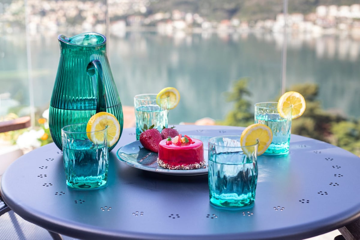 A round table is set for refreshments, featuring a teal pitcher and four matching glasses with lemon slices. A plate holds a pink dessert garnished with fresh strawberries, all against a backdrop of a serene bay view reflecting the surrounding landscape.
