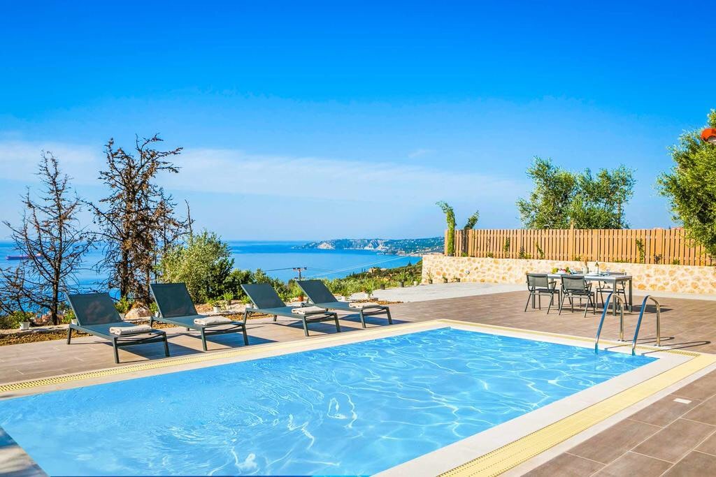 A private pool is centered within a sunny outdoor space, surrounded by comfortable lounge chairs. Clear views of Lourdas Bay and the Greater Ionian Sea can be seen in the background, while a dining area is positioned nearby, framed by well-maintained greenery.