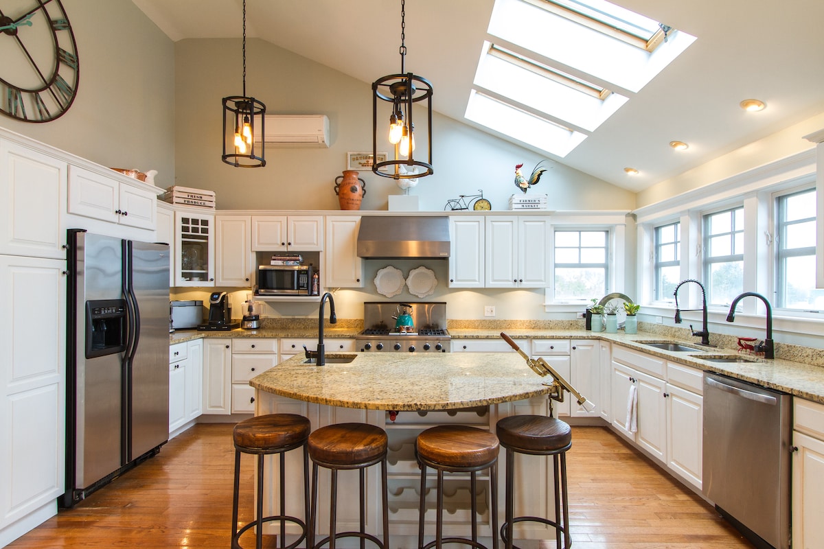 A spacious kitchen is characterized by white cabinetry and granite countertops. Skylights provide ample natural light, while pendant lights hang over a central island with seating. Appliances are modern and stainless steel, complementing the inviting atmosphere of the room.