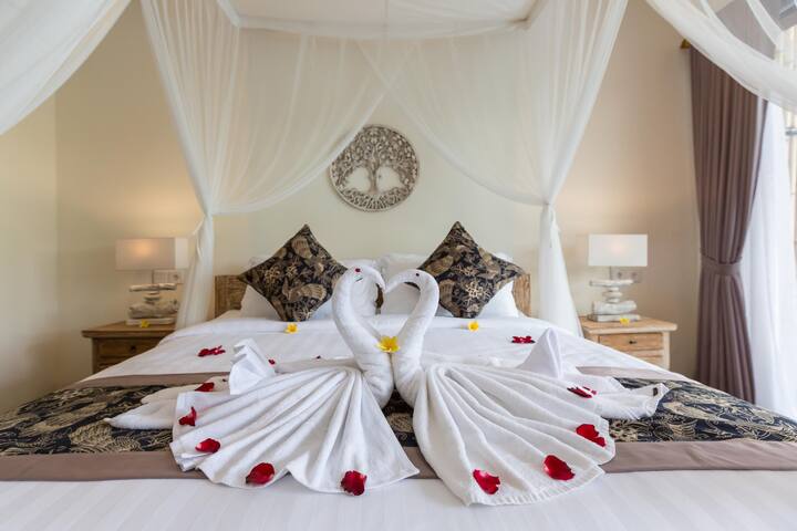 Романтична спалня с легло с балдахин, покрито с прозрачна бяла тъкан, украсено с декоративни лебеди и цветя.  Мекото осветление и топлите тонове създават луксозна и интимна атмосфера.