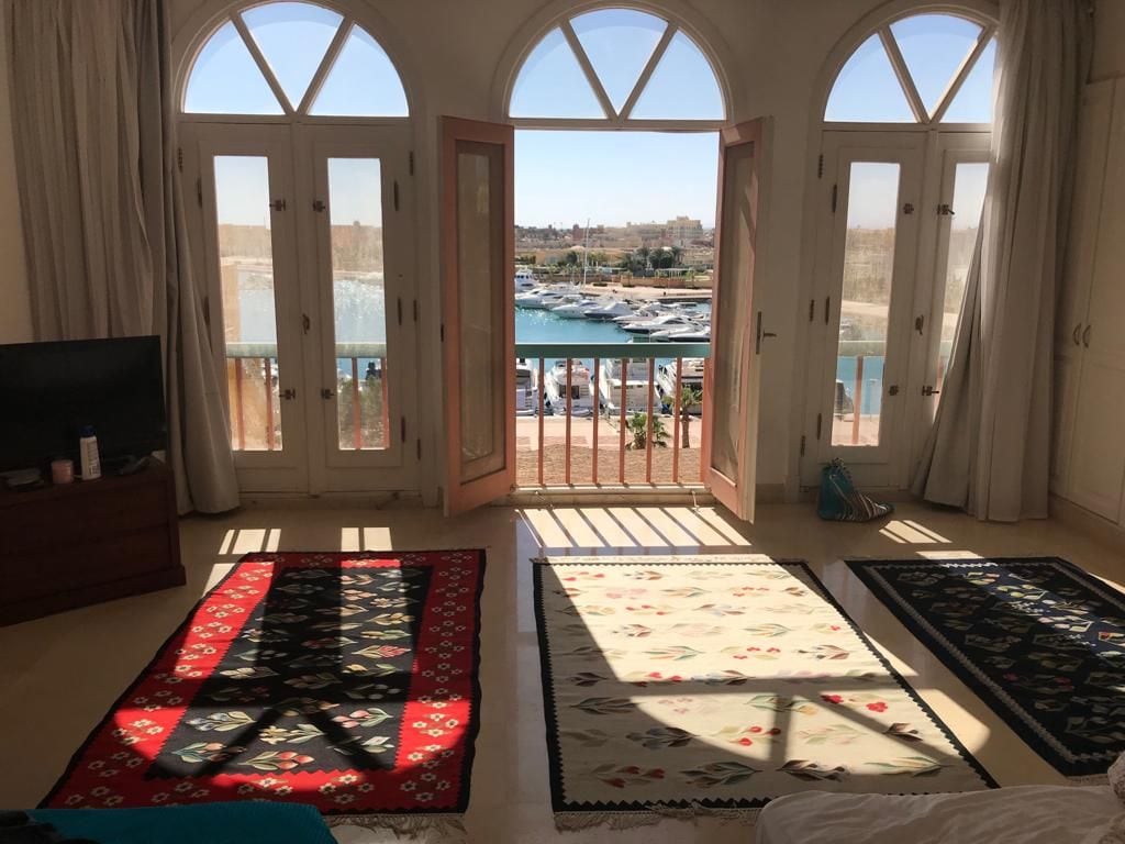 A spacious room features large arched windows with views of the marina and boats. Two area rugs, one red and one patterned, are laid on the floor, with sunlight casting shadows. A wooden dresser is positioned against the wall, enhancing the room's bright and airy atmosphere.