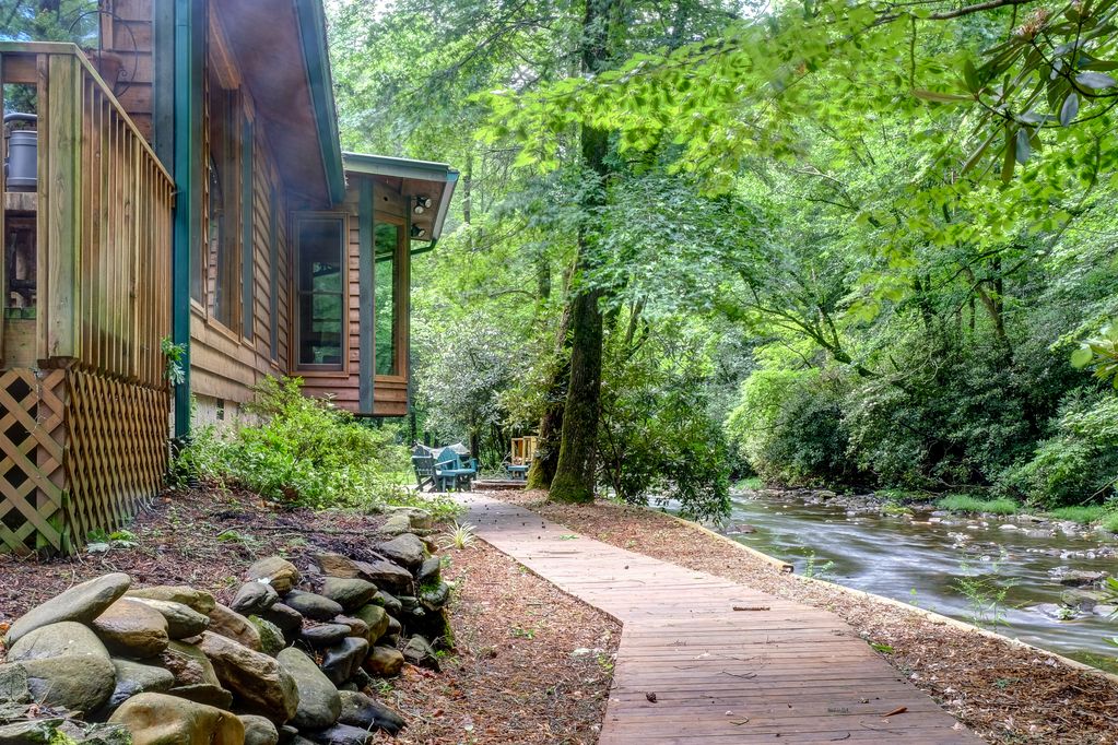 A natural pathway leads to the edge of a gently flowing creek. Stone landscaping borders the walkway, which is flanked by lush greenery and trees. The cabin's wooden exterior is visible nearby, with outdoor seating arranged for relaxation along the water's edge.