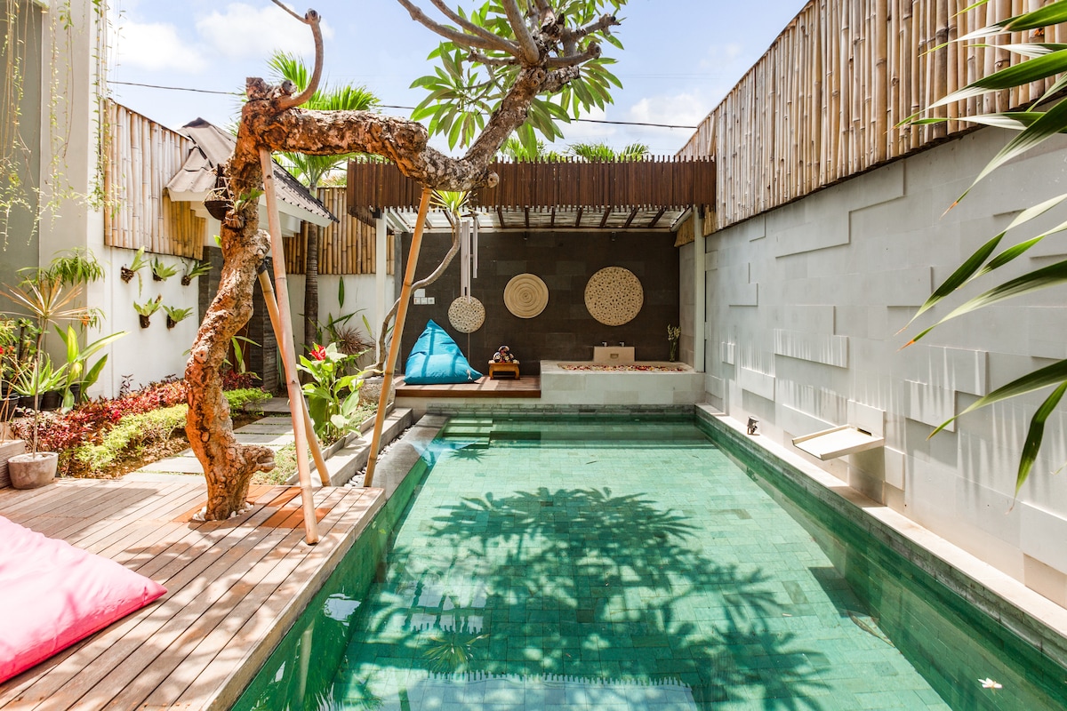 A tranquil outdoor area features a swimming pool surrounded by lush greenery and wooden decking. A unique tree sculpture stands beside the pool, while a decorative wall showcases natural textures. A relaxing jacuzzi is also present, enhancing the serene ambiance.
