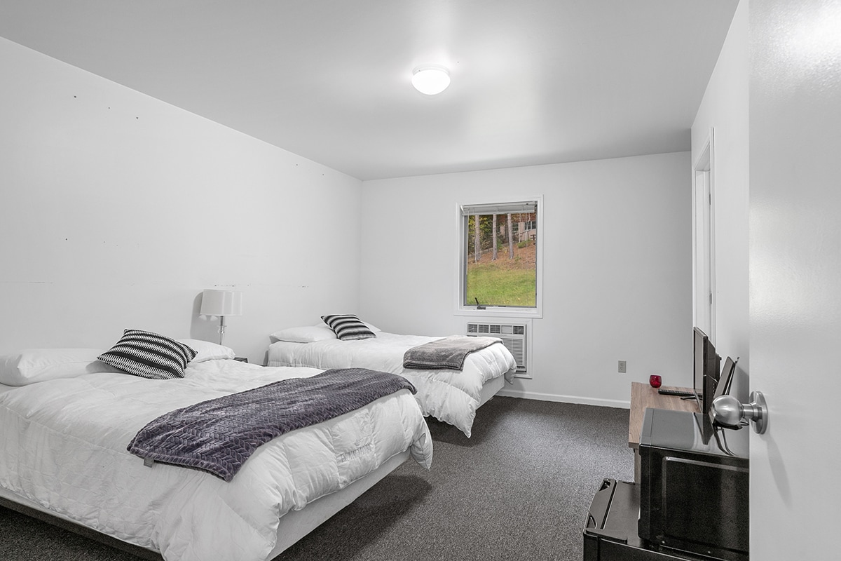A spacious hotel room contains two Full beds dressed in white linens, complemented by dark striped throw pillows. A window faces the outdoors, allowing natural light to enter. A flat screen TV and a small refrigerator are positioned near the entrance.