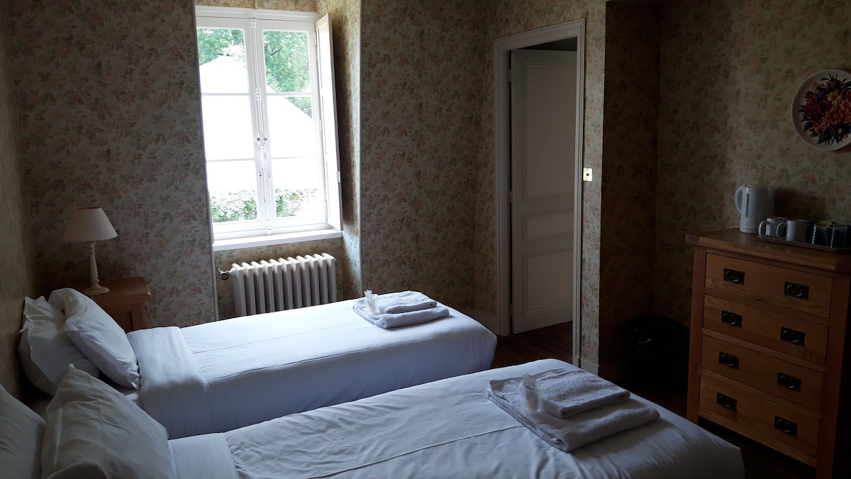 A cozy room featuring two single beds with neatly arranged white linens. A window allows natural light to enter, complemented by floral wallpaper. A wooden dresser is positioned against one wall, while a kettle and cups are visible on a nearby table.