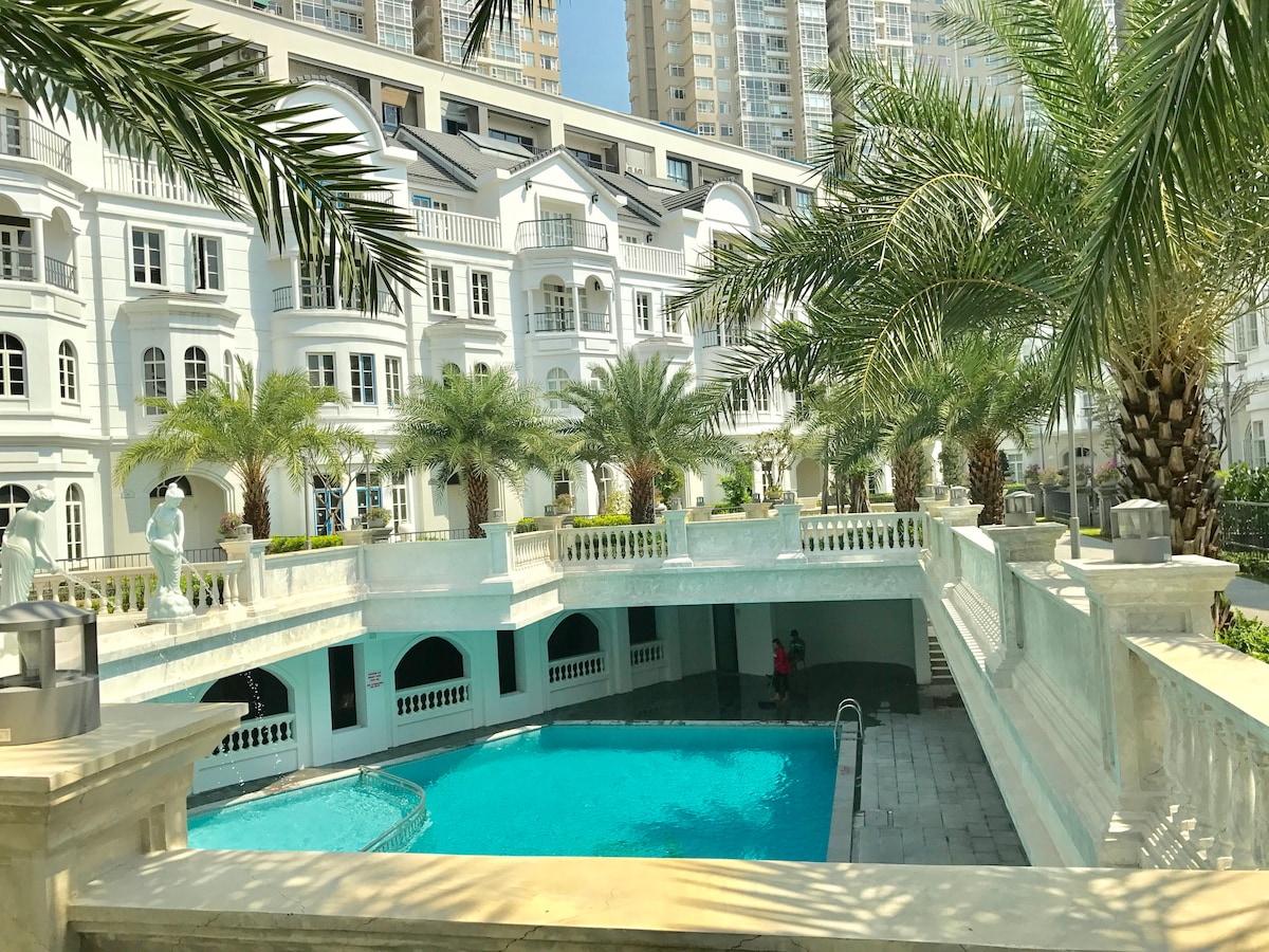 A serene swimming pool is framed by palm trees and elegant white architecture. The softly flowing water is highlighted within a well-maintained courtyard, creating a relaxing ambiance. Sunlight reflects off the pool's surface, enhancing the inviting atmosphere of the surrounding greenery.