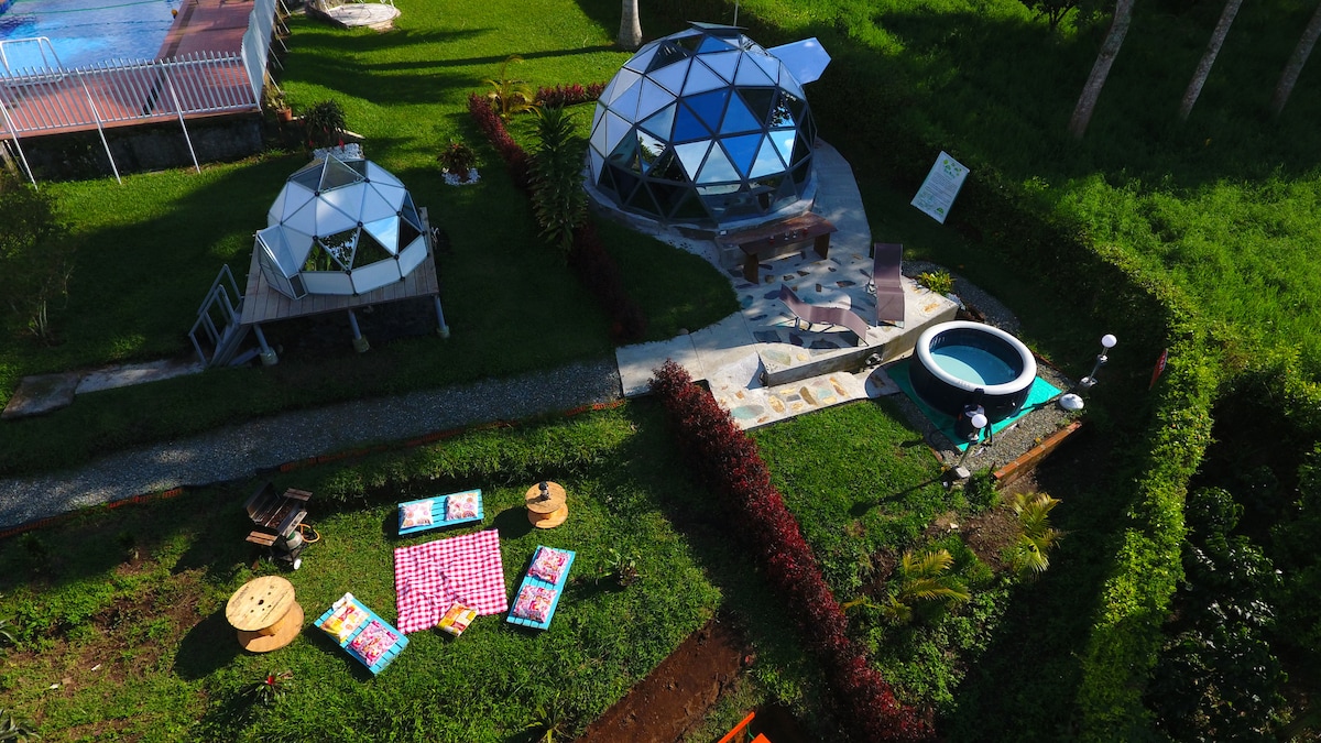 Two geodesic structures are situated in a lush green area, featuring large glass sections that enhance the natural surroundings. A private jacuzzi with a vibrant blue finish is nearby, and a seating area is arranged with colorful cushions on a grassy patch, inviting relaxation.