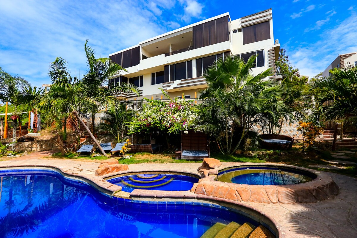 The exterior view showcases a modern villa with a spacious balcony and large windows. A vibrant pool area features a primary pool and a small jacuzzi surrounded by tropical plants. Stone pathways lead to lounge chairs under tall palm trees, creating a relaxing outdoor space.