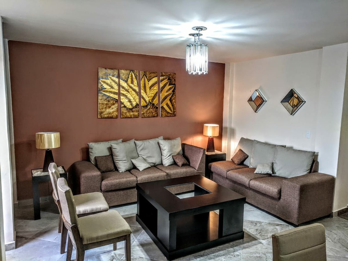 A spacious living area features a cozy arrangement of brown sofas, complemented by decorative cushions. A dark coffee table with a glass top sits in the center, while two stylish lamps provide soft lighting. Artwork adorns the walls, enhancing the ambiance.