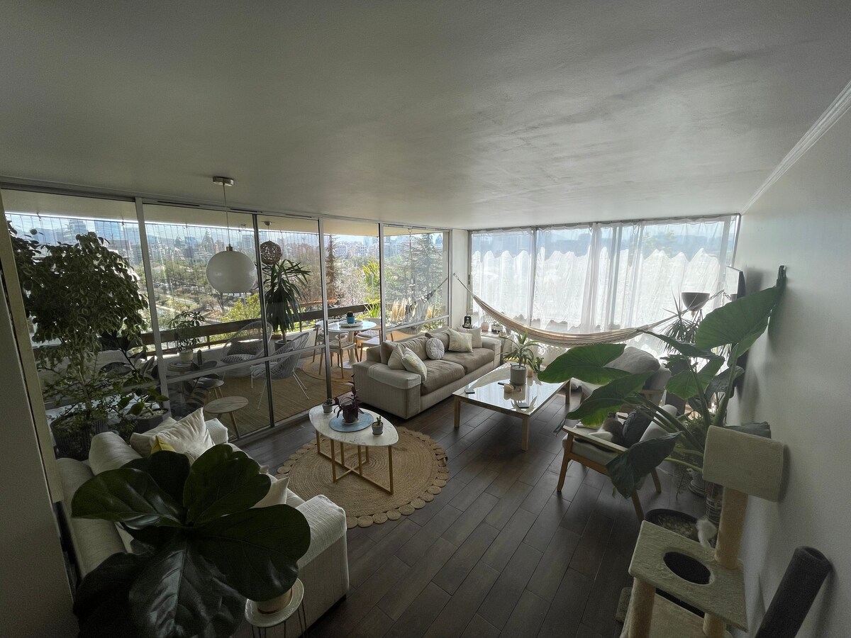 A spacious living room features large windows that provide ample natural light and a view of the outdoors. A sectional sofa and coffee table are arranged around a central area. Various plants enhance the atmosphere, while a hammock adds a relaxed touch to the seating area.