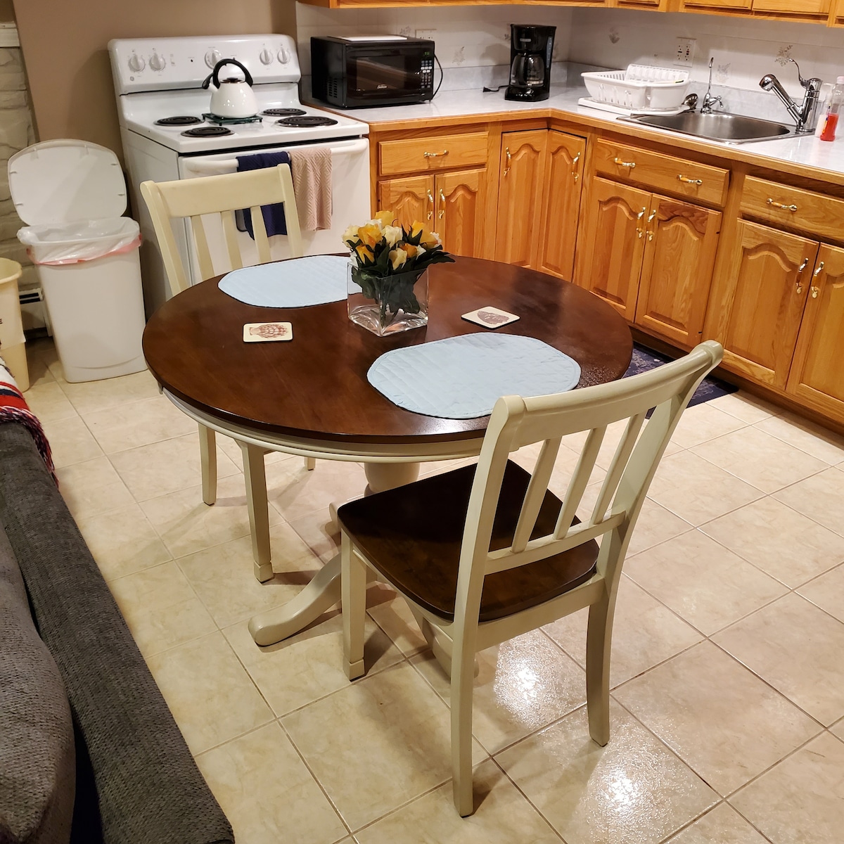 A round dining table with two white chairs is positioned in the kitchen area. A small vase of flowers is centered on the table, complemented by two blue placemats. Kitchen appliances such as a microwave, coffee maker, and stove are visible in the background, with wooden cabinetry providing storage.