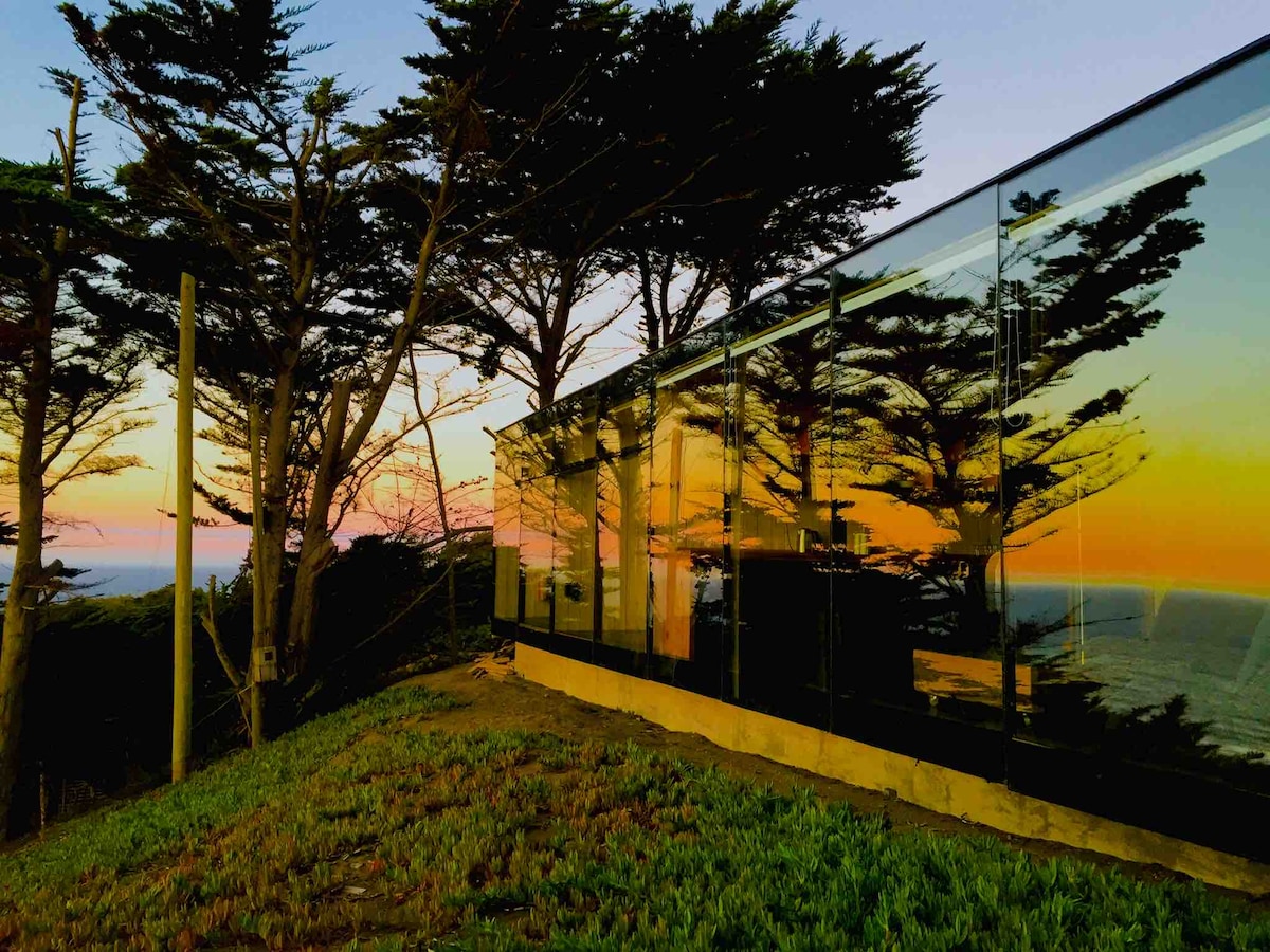 A modern house is positioned along the hillside, featuring large glass walls that reflect the surrounding trees and sunset colors. The foreground includes a lush green area, while the background offers views of the ocean, enhancing the home's integration with nature.