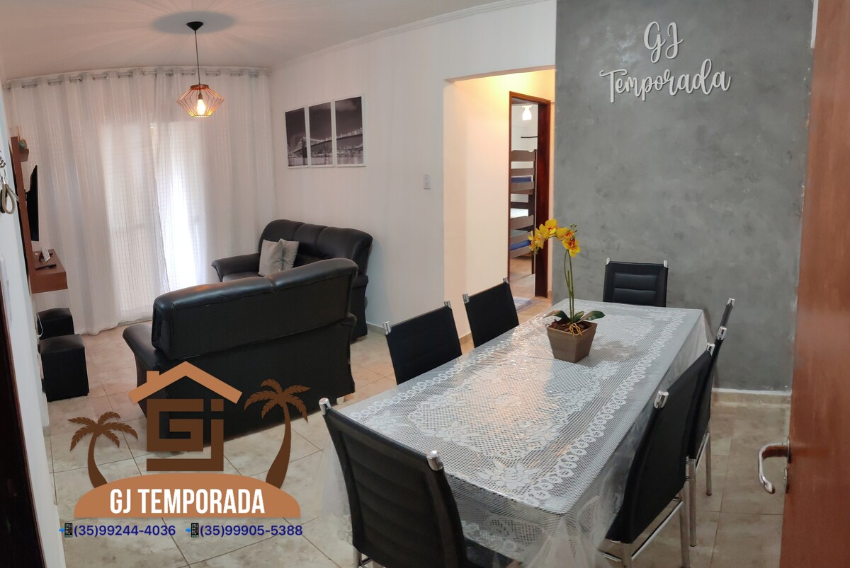 The image displays a spacious living and dining area featuring a dark sofa and a glass dining table surrounded by six chairs. A vase with yellow flowers is placed at the center of the table. Natural light filters through sheer curtains in the background.