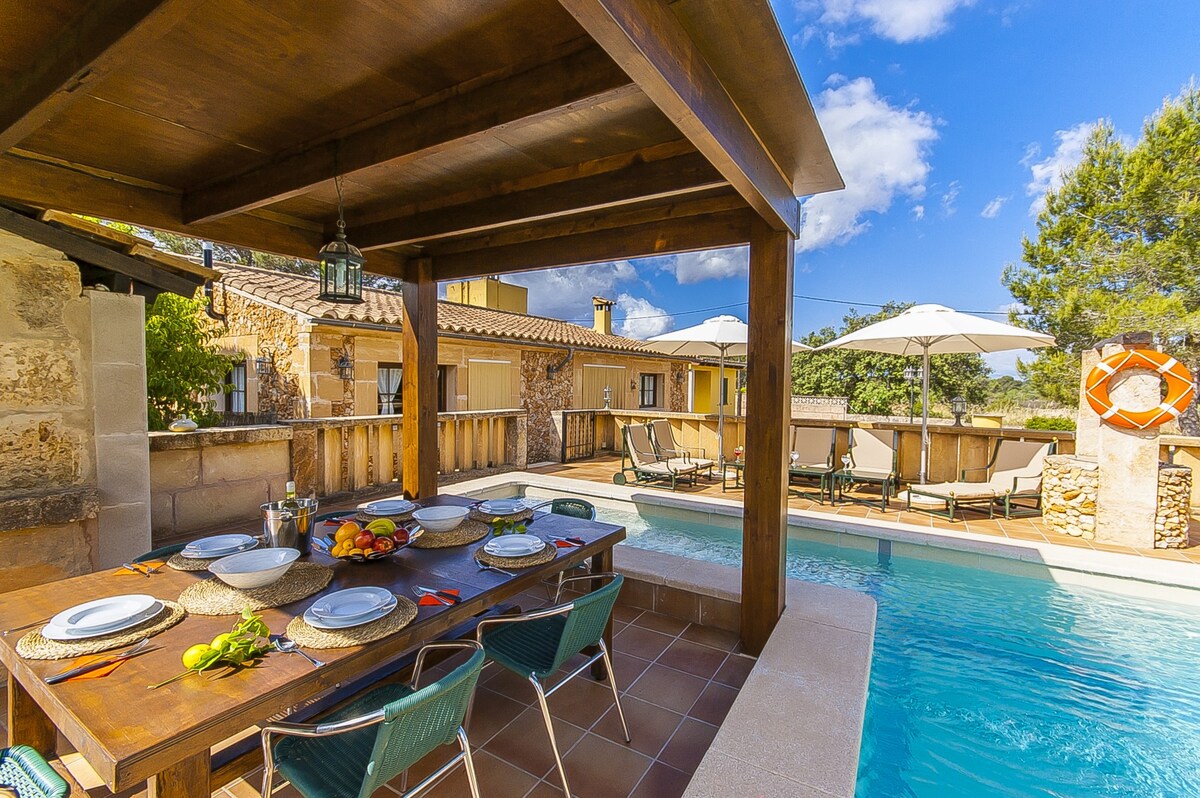 An outdoor dining area features a wooden table set with dishes and fresh fruit. Enclosed by a roofed structure, the space offers a pool and sun loungers under large umbrellas in the background. The sunlit setting is complemented by greenery and blue skies.
