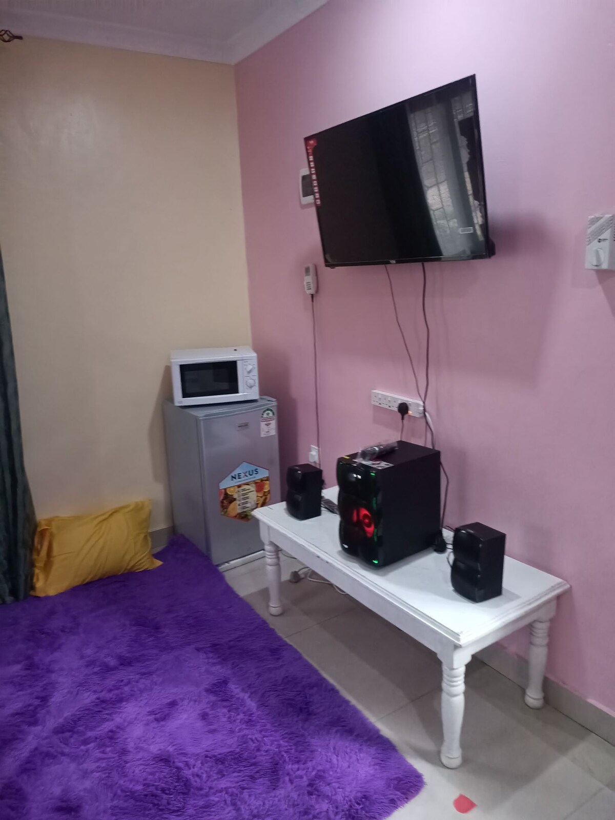 A small living area features a wall-mounted television above a white table. A computer tower and speakers are positioned on the table. Adjacent, a compact refrigerator and microwave are accessible. The floor is covered with a plush purple rug, complemented by an orange cushion.