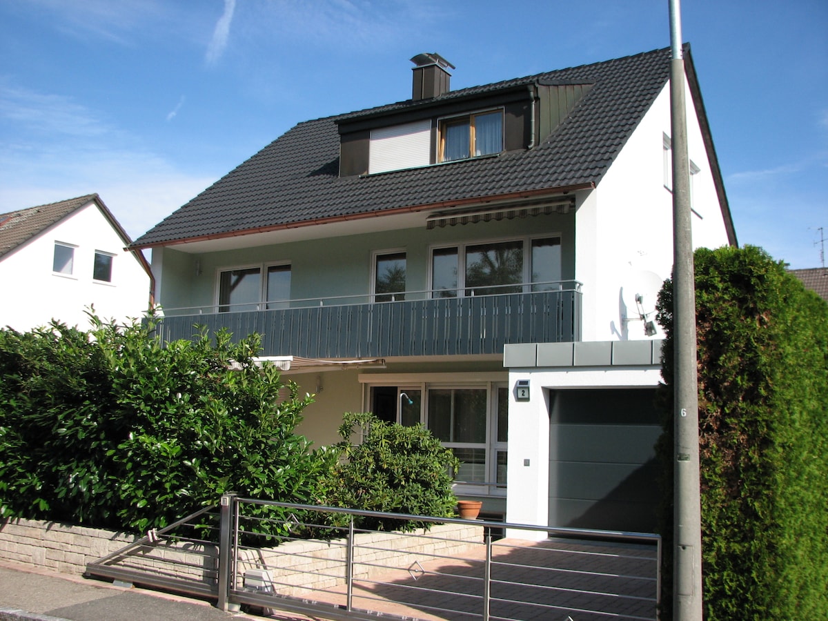 A multi-story house is shown, featuring a combination of light blue and white exterior walls. A balcony is present on the upper floor, with surrounding greenery providing some privacy. The driveway leads to a garage, and the entrance is visible through glass doors.