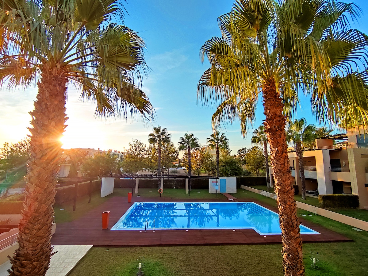 The image captures a serene swimming pool surrounded by palm trees, with a wooden deck offering ample space. Sunlight filters through the palms, creating a warm glow over the pool's clear water. Lush green grass completes the peaceful atmosphere of the outdoor area.