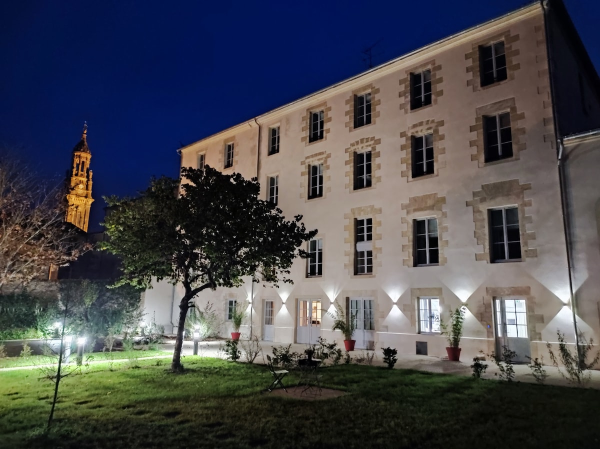The exterior of the residence is showcased at night, highlighting the warm illumination from ground lights. The building features a light-colored facade with multiple windows, while a clock tower is visible in the background. The garden area contains greenery and flower pots, adding a serene ambiance.