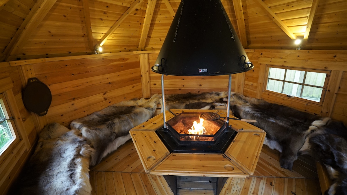 The interior of the BBQ hut features warm wooden walls and a central fireplace, surrounded by benches covered in soft fur. The area is illuminated by subtle light fixtures, creating a cozy setting for gatherings, with windows providing views of the surrounding garden.