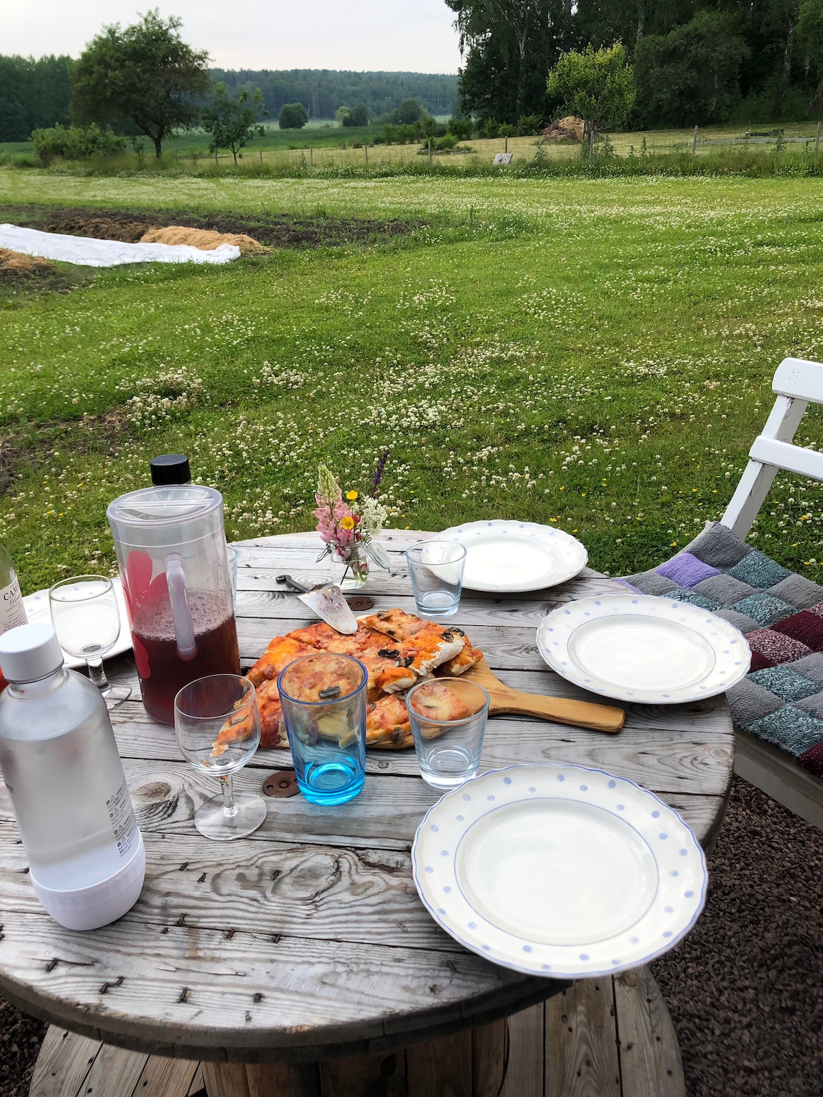 A rustic wooden table is set outdoors, featuring several plates and glasses. A colorful pizza on a wooden board serves as the centerpiece, complemented by a pitcher and a small bouquet of flowers. An expansive green field borders the scene, providing a serene backdrop.