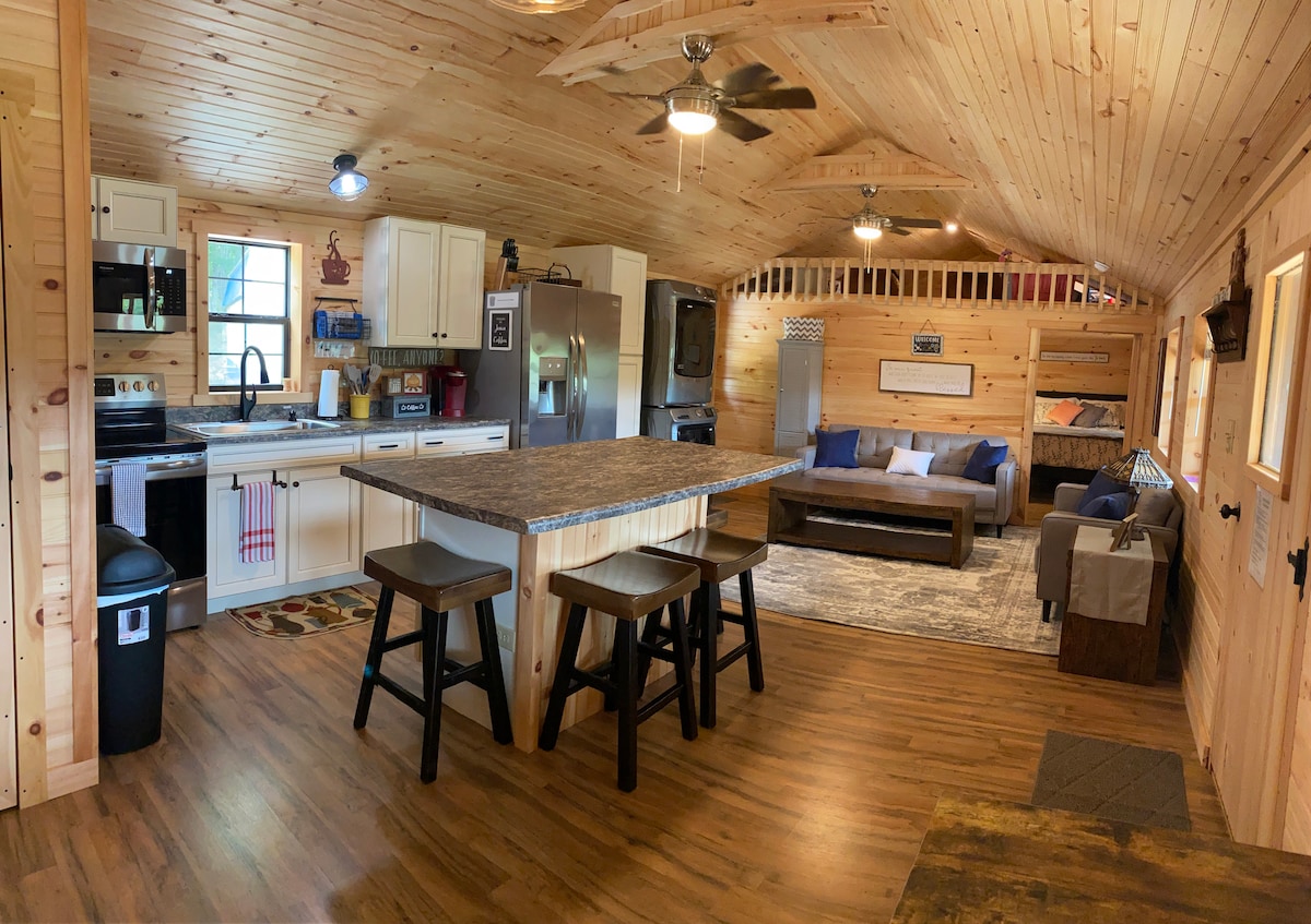 An open-plan living space features a full kitchen with wood cabinetry and modern appliances, including a refrigerator and a range. A large island with seating complements the dining area. The cozy living area includes a sofa and decorative rug, all set beneath wooden ceilings with ceiling fans.
