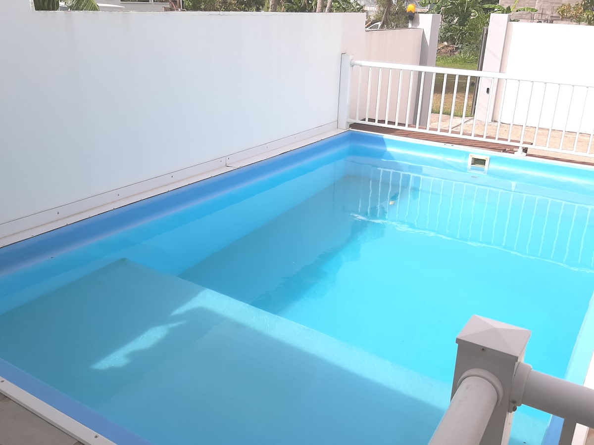 A private pool is visible, featuring clear blue water and surrounded by a white fence. The pool area is well-lit, emphasizing the tranquil ambiance. Sunlight reflects off the surface of the water, creating a serene atmosphere.