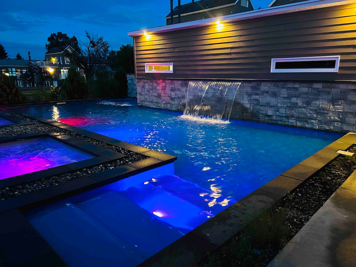 A well-lit swimming pool is featured, showcasing a gentle waterfall and illuminated in shades of blue and purple. The surrounding area includes stone accents and landscaping, creating a serene atmosphere. The pool is designed for relaxation and enjoyment.