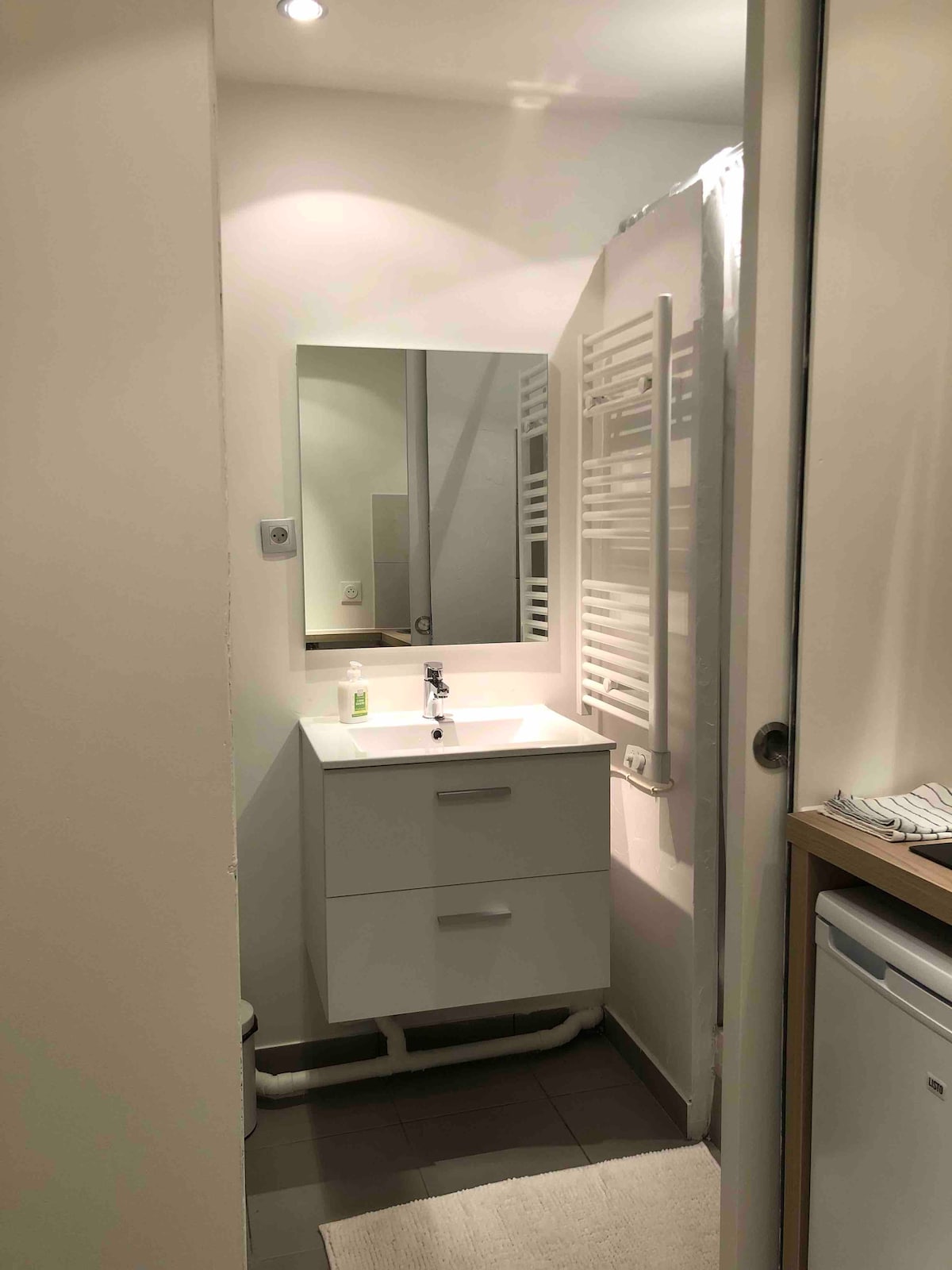 A compact bathroom features a modern white vanity with a sink and storage drawers. A large mirror reflects the space, enhancing the bright, inviting atmosphere. A towel radiator is mounted on the wall, and simple, neutral tiles cover the floor.