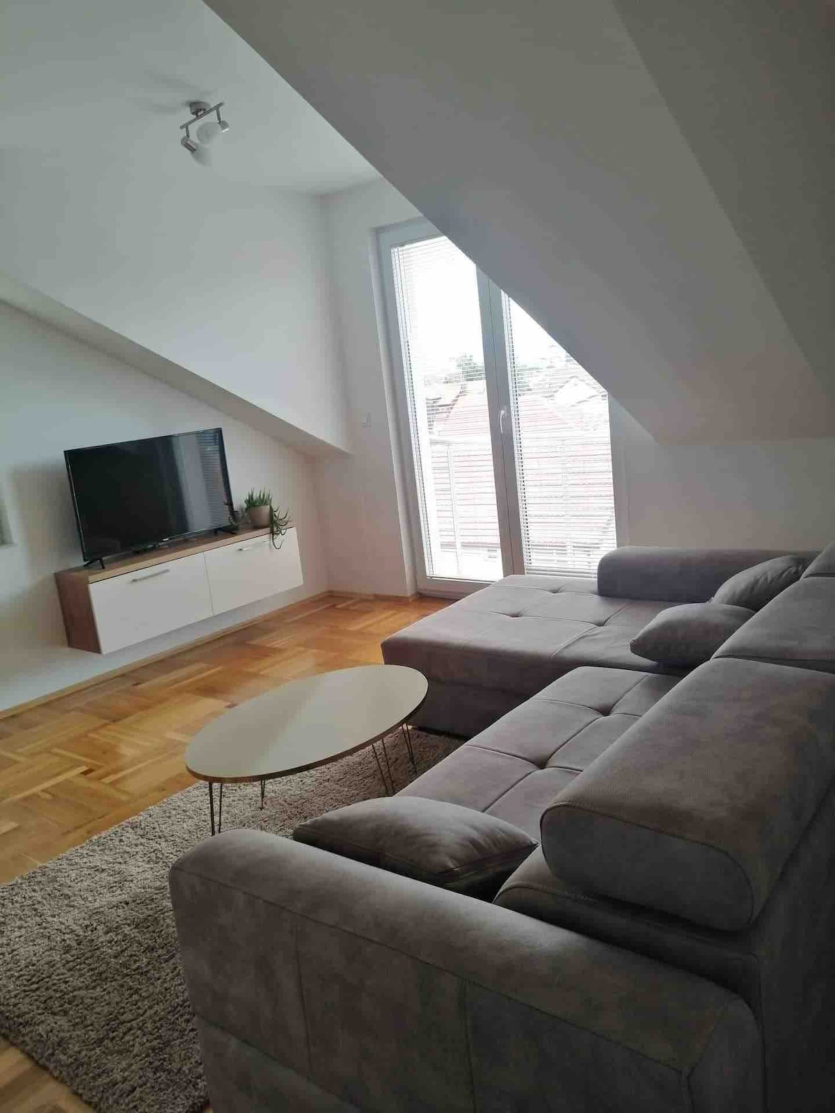 A cozy living area features a sectional sofa in a soft grey hue, complemented by a small round coffee table. A flat-screen TV is mounted on the wall above a sleek media console, and natural light filters in through a glass door leading to a balcony.