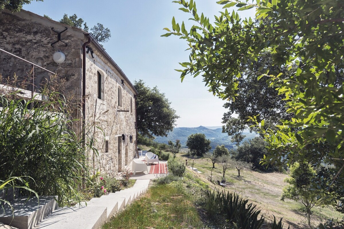 A stone farmhouse is surrounded by lush greenery and rolling hills. The outdoor area features a staircase leading to a terraced space, with seating visible under a tree. The landscape extends into the distance, showcasing valleys and hills under a clear sky.