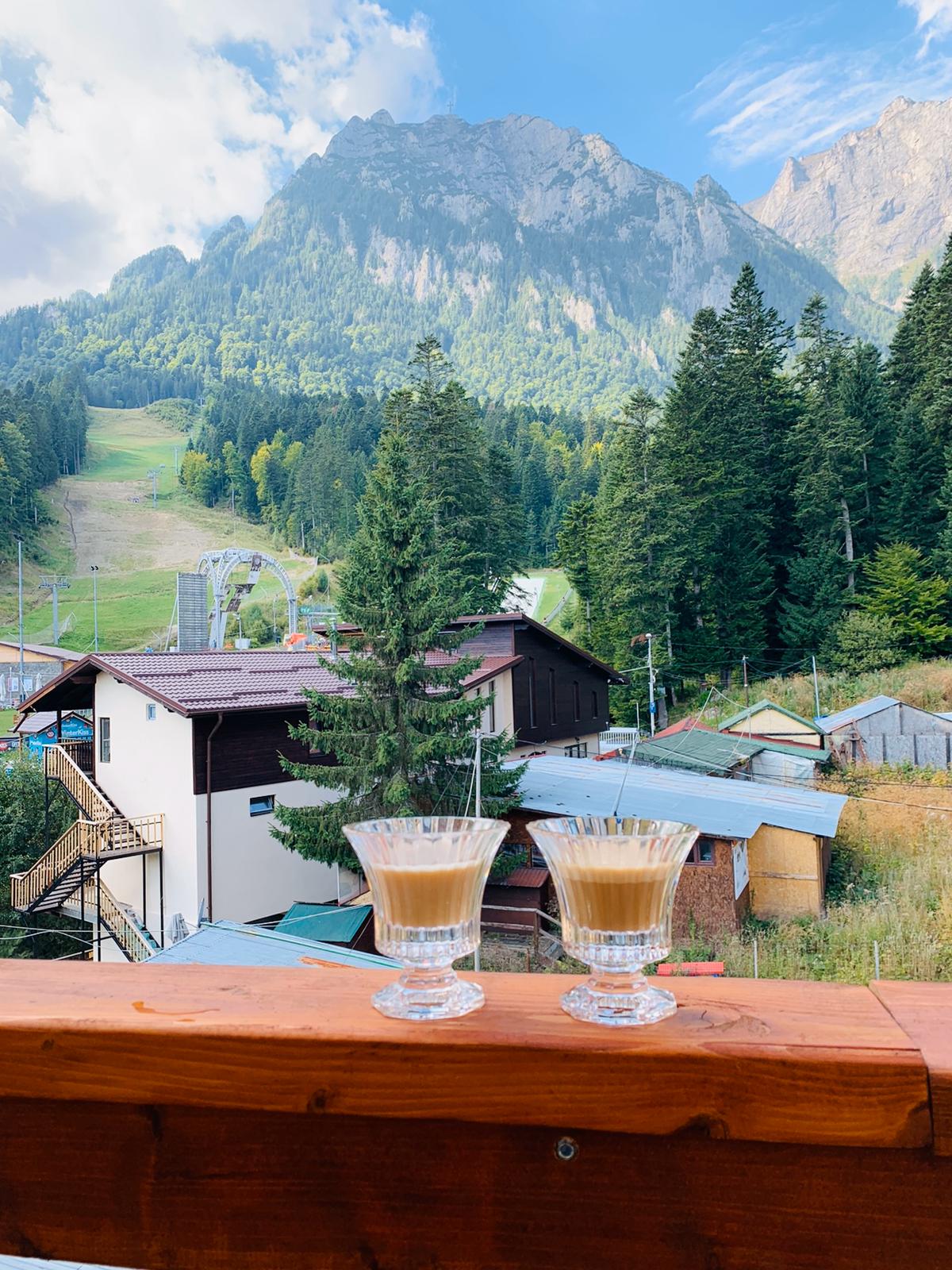 Two glasses of coffee are placed on a wooden balcony railing, overlooking a picturesque landscape. Towering mountains loom in the background, with lush trees and ski slopes visible beneath a partially cloudy sky.