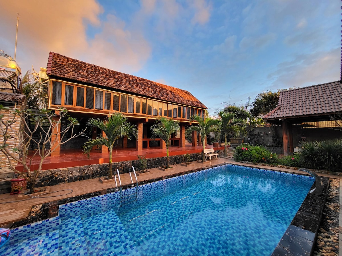 An inviting outdoor pool area is framed by palm trees and colorful flowers. A traditional wooden house with large windows overlooks the pool, reflecting a serene environment. The pool features steps leading in, with a tiled surface aiding in visual contrast against the surrounding greenery.