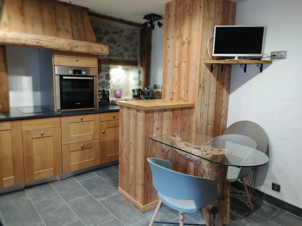 A well-equipped kitchen features wooden cabinetry and stone accents. A modern glass dining table is complemented by two minimalist chairs. An oven and microwave are built into the cabinetry, while a TV is mounted on the wall, enhancing the functional design of the space.