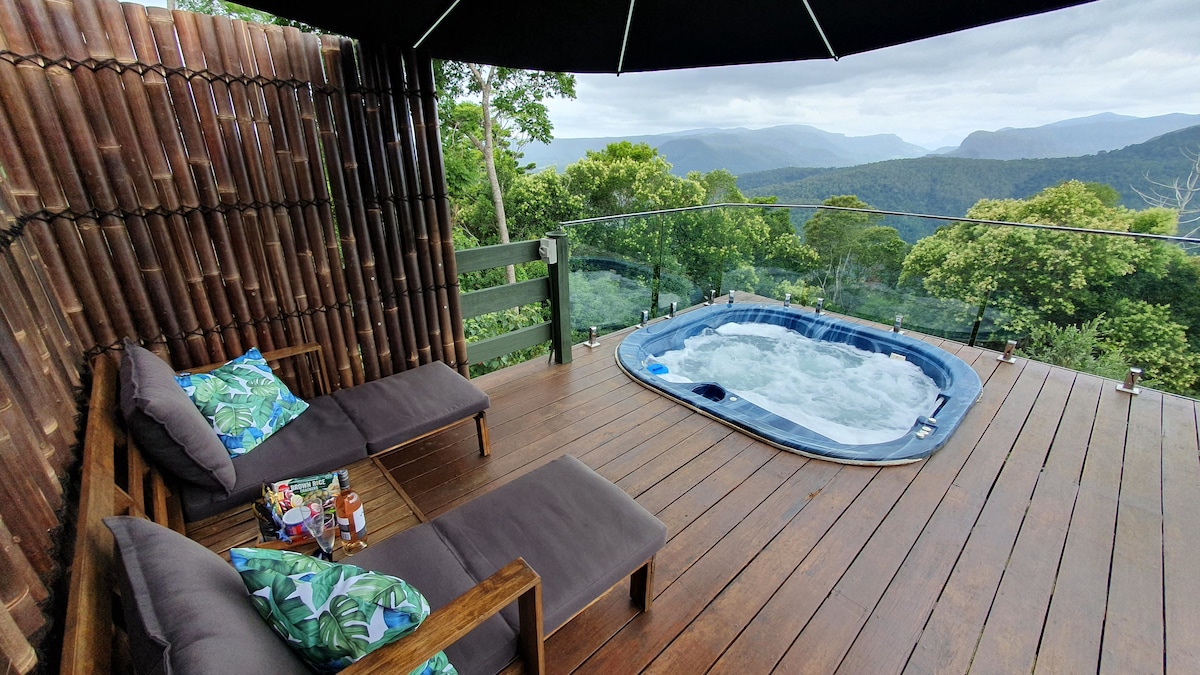 A private outdoor space features a heated spa and two sunbeds, surrounded by a wooden deck. Lush greenery and mountain views are visible in the background, with a shaded umbrella providing comfort. Decorative cushions add a touch of color to the seating.