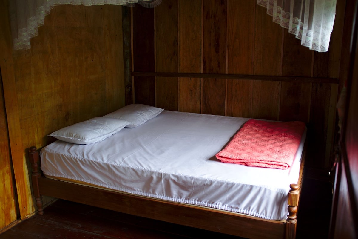 A simple sleeping area features a wooden bed with a white mattress and two pillows. A red blanket is neatly placed at the foot of the bed. Natural wood walls create a warm atmosphere, while soft light filters in from a nearby window covered by lace curtains.