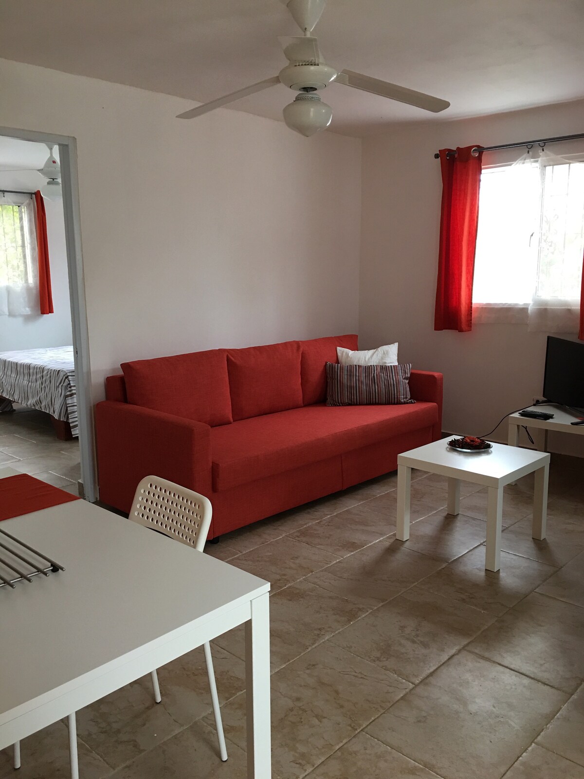 A cozy living area features a bright orange sofa and a small white coffee table. Natural light streams through the large window, adorned with light curtains. A dining table with white chairs is visible, and a television is positioned on a stand near the wall.