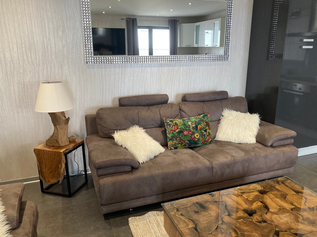A cozy living area features two brown sofas with decorative cushions, paired with a wooden side table and a unique glass coffee table. Soft lighting from a lamp enhances the relaxed ambiance, while a large mirror reflects the space.