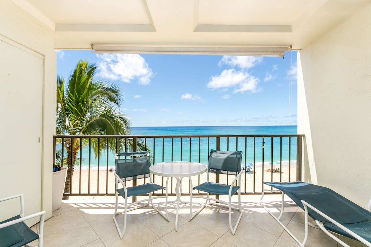 A private lanai offers a table and several lounge chairs with ocean views. The tropical landscape includes a palm tree, and the vibrant blue ocean extends seamlessly to the horizon. Natural light illuminates the space, enhancing the outdoor atmosphere.