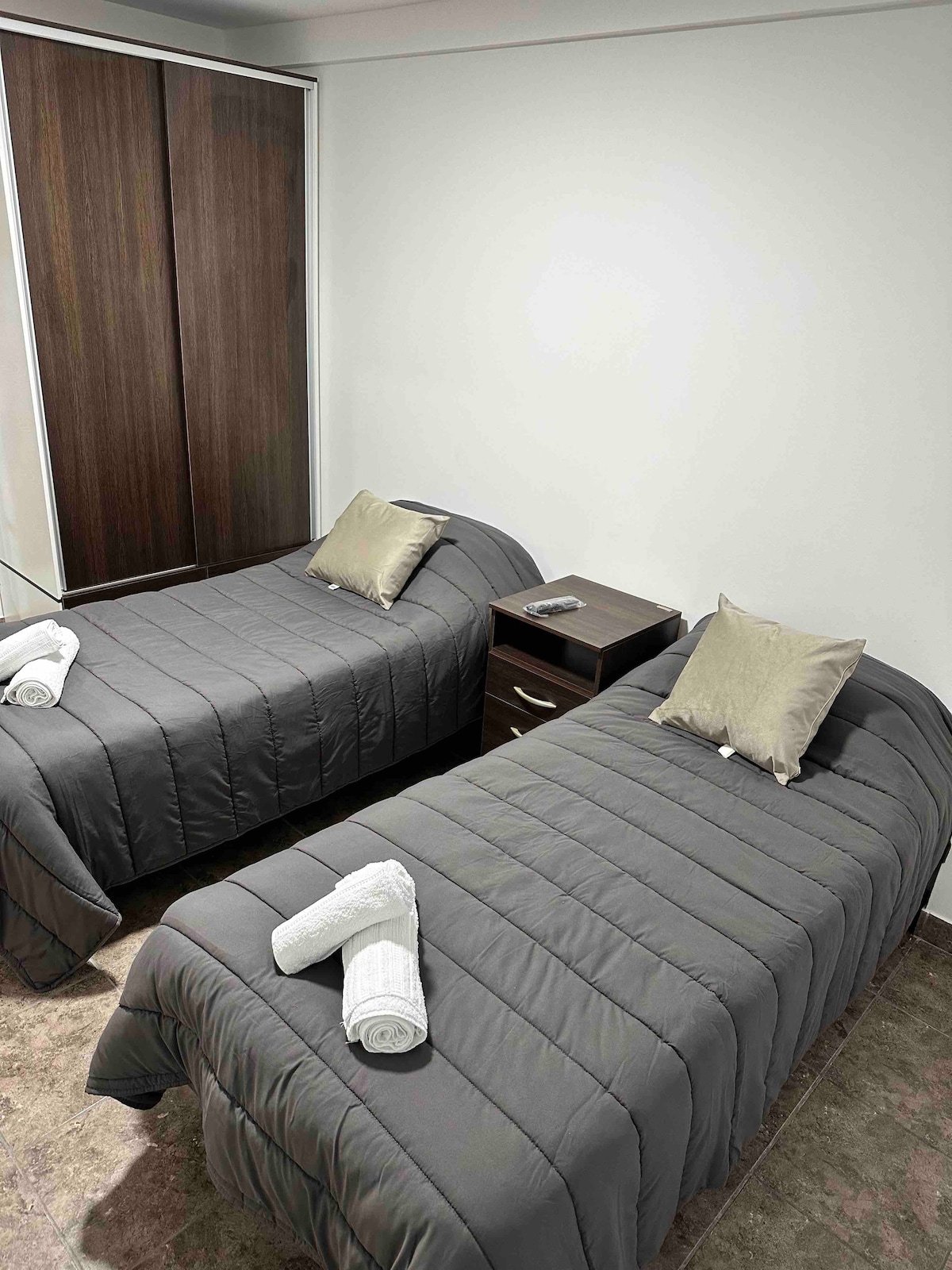 A functional sleeping area is presented with two individual beds, each adorned with gray bedding and decorative pillows. A small nightstand is placed between the beds, featuring a notepad and a pen. Soft white towels are neatly arranged at the foot of each bed.